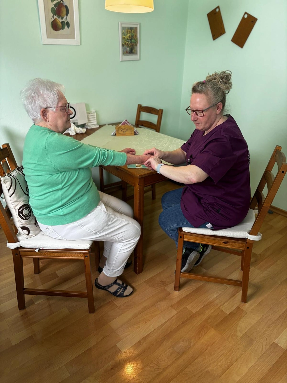Pflegedienst Döring - Ambulanten Pflegedienstes - Eine ältere Frau in einem grünen Pullover sitzt an einem Holztisch, während eine medizinische Fachkraft in violetter Arbeitskleidung ihre Hand untersucht. Sie befinden sich in einem hell erleuchteten Raum mit hellgrünen Wänden und Holzboden.